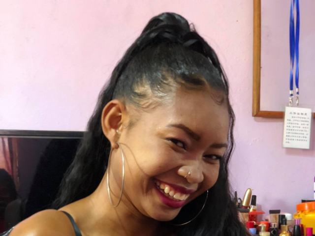 Chelina with black ponytail, smiling, hoop earrings, nose ring, and pink lipstick. Her dark skin and red lipstick contrast with her white tank top. Her tits are small.