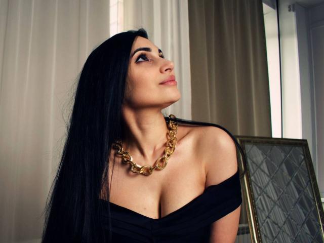 AllaYertlyi with long black hair, looking up, wearing a black off-shoulder top, gold chain necklace, showing her boobs, in a room with a mirror and curtains.