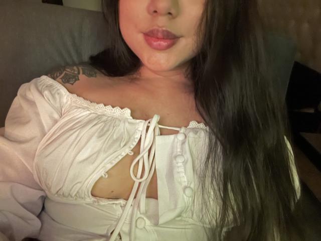 SteffanyTaylor with long black hair, wearing a white off-shoulder top, exposing her tits, with a tattoo on her left shoulder, and a small amount of cum on her chin.