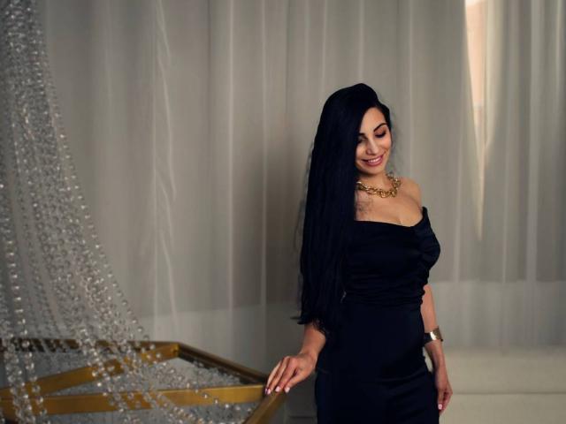 AllaYertlyi with long black hair, smiling, wearing an off-shoulder black dress, gold necklace, and bracelet, standing by a beaded curtain, holding her ass with her right hand.