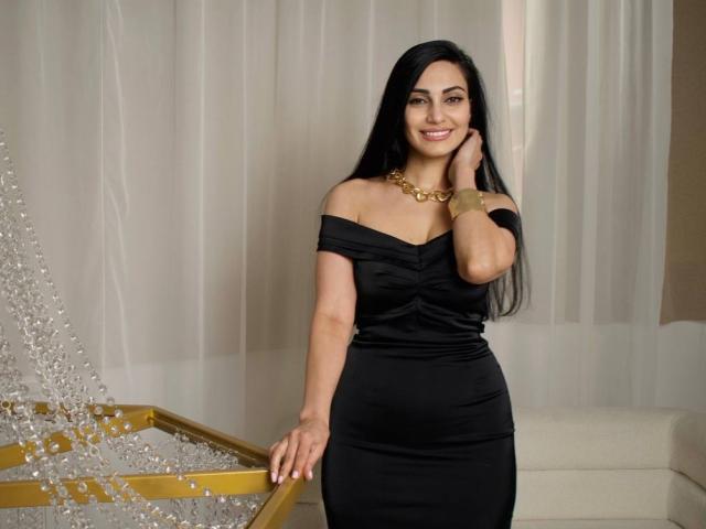 AllaYertlyi with long black hair, off-shoulder black dress, gold necklace, smiling, hand on cheek, standing by a gold and silver Christmas tree. Boobs visible under dress.
