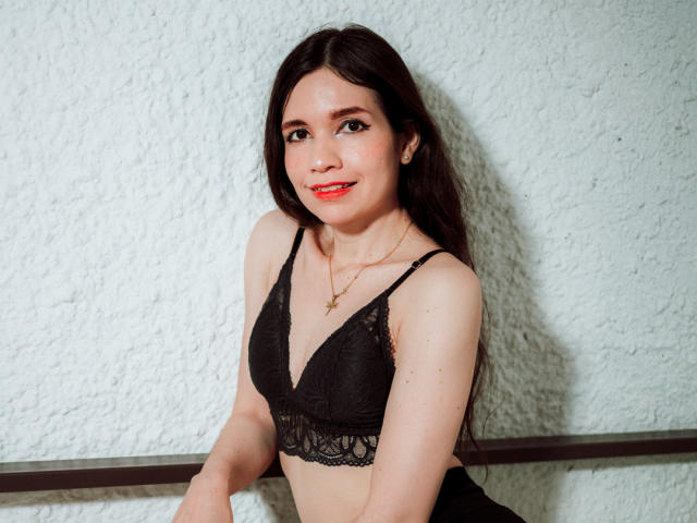 SofiArias with long dark hair, wearing a black lace bra, smiling with red lipstick, small tits, light skin, and a gold necklace, leaning against a white textured wall.