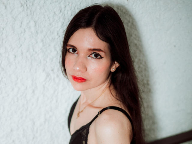 SofiArias a de longs cheveux bruns foncés, du rouge à lèvres, et porte un haut en dentelle noire qui révèle ses petites seins, avec un léger sourire, et un mur blanc texturé en arrière-plan..