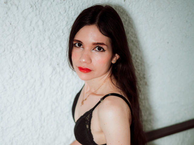 SofiArias with long dark hair, red lipstick, black lace bra, small tits, fair skin, standing against white textured wall, looking at camera with slight smile, gold necklace.