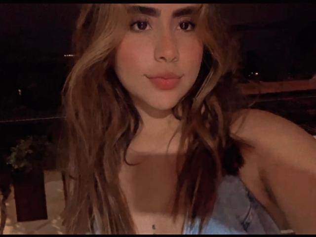 OlivaStellas, con cabello castaño ondulado, mejillas rosadas y una pequeña peca debajo de los senos, vestida con una blusa azul, sonriendo suavemente en una foto con poca luz y tonos cálidos..