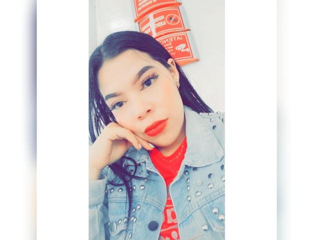 AdrianaPaul, con cabello negro largo, lápiz labial rojo y una chaqueta vaquera azul adornada con tachuelas, recostando la cabeza en su mano, vistiendo una camisa roja, luciendo linda y segura de sí misma..