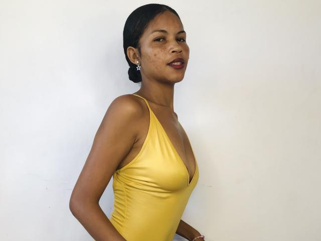 CharlineMagik with dark skin, wearing a yellow, deep V-neck, one-shoulder dress, showing her small tits and slim figure, standing against a white wall, looking at the camera.