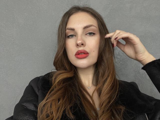LexxiJoy with long brown hair, red lipstick, and a black jacket, has a hand in her hair, showing red nails. She has a small mole under her right eye.