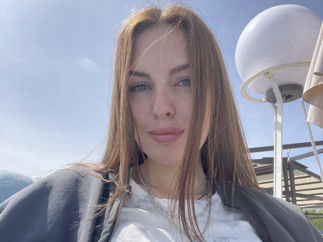 LexxiJoy has straight brown hair, blue eyes, and a neutral expression. She's wearing a white shirt and gray hoodie with a silver chain, standing outdoors in sunlight.