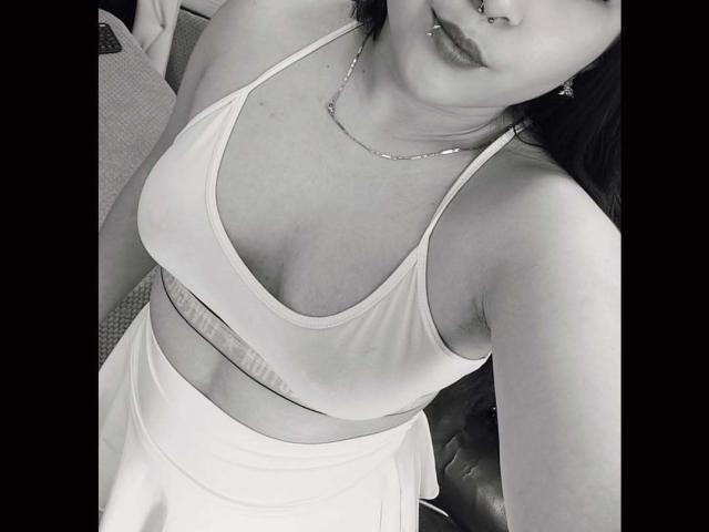 ElinaX with medium tits in a white sports bra and skirt, silver necklace, nose ring, and dark hair, looking down at the camera. Monochrome selfie.