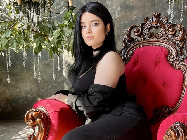 HottieShy with long black hair, pale skin, and sharp eyeliner sits on a red, ornate wooden chair, wearing a black top and pants with a black jacket off-shoulder.