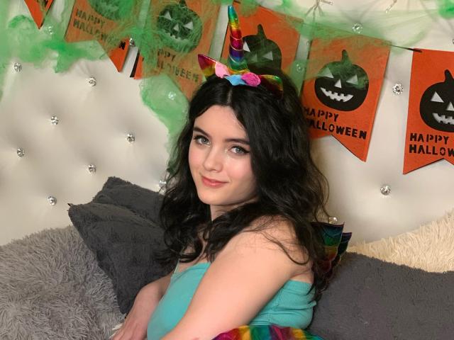 HottieShy with long black hair, colorful horn, and green eyes, wearing a blue top, sitting on a gray and fluffy pillow, smiling at the camera. HottieShy has tits.