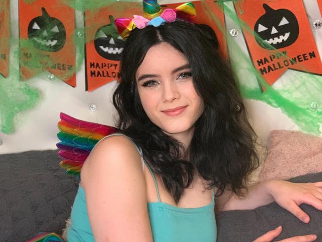 HottieShy con cabello negro ondulado, diadema de arcoíris, alas coloridas y top color turquesa, sonriendo a la cámara, mostrando sus pequeños pechos, linda y sexy..