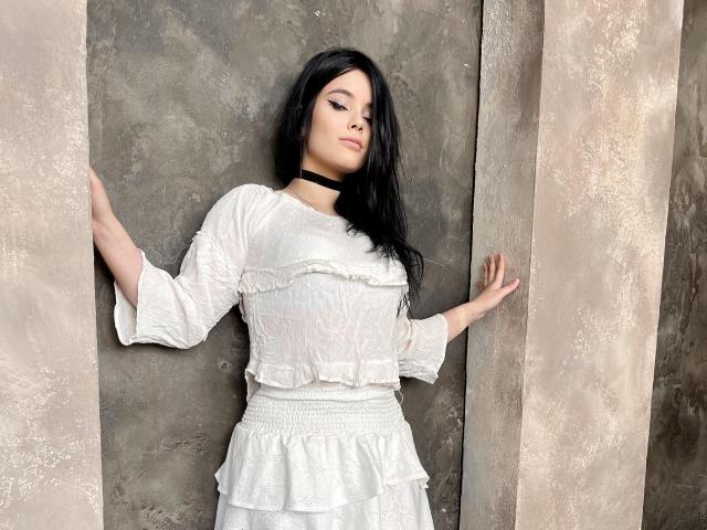 HottieShy with black hair, white dress, and black choker, standing against rough concrete, hands on walls, eyes closed, confident, sexy, subtle makeup, medium-sized tits, soft light.