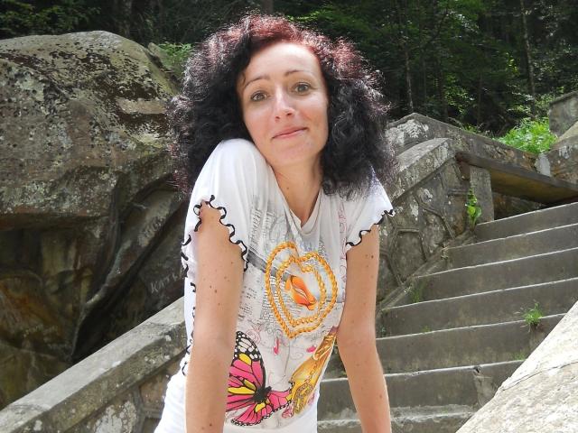 Pussyxxx, aantrekkelijk met krullend zwart haar, een wit shirt met een kleurrijke vlinder, leunt tegen stenen trappen, een groen bos op de achtergrond, glimlacht zachtjes, lichte huid, gemiddelde borsten, natuurlijk licht..