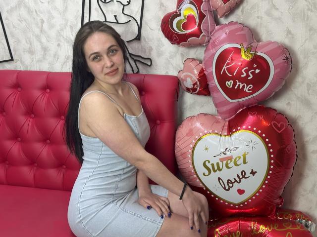 IziPaterson with long brown hair, wearing a light blue dress, sitting on a pink tufted couch, next to shiny pink heart balloons, one labeled "Kiss me" and "Sweet love".