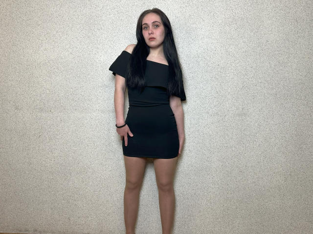 IziPaterson stands with long black hair, wearing a black off-shoulder dress, showing her bare ass, and a black wristband, against a plain, textured wall.
