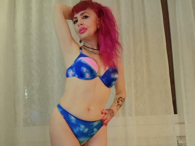 HotAdelayde69 with pink hair, blue tie-dye bikini, small tits, tattoo on right arm, hand on head, standing, white curtains, pale skin, red lipstick, confident pose.