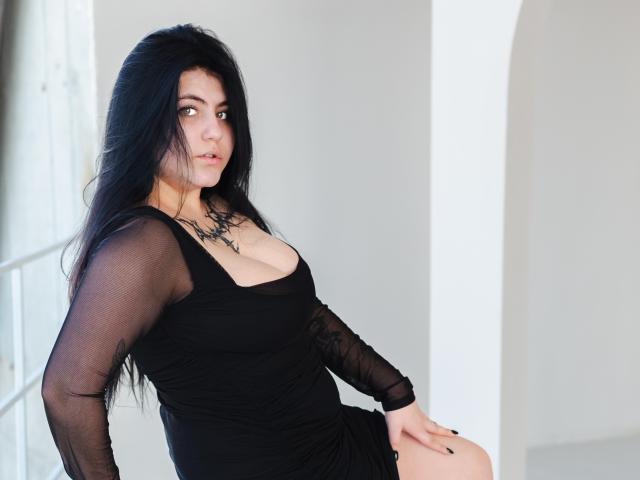 NanaDaizy mit langen, schwarzen Haaren, trägt ein durchsichtiges schwarzes Kleid, das ihre Brüste zeigt, sitzt mit einer Hand auf ihrem Oberschenkel, blickt zur Seite, im Hintergrund eine weiße Wand..