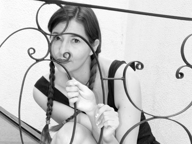AntonnellaS with pigtails, black dress, holding metal railing with swirls, sucking on metal, black and white, sitting, looking at camera, soft expression, curly metal, textured wall.
