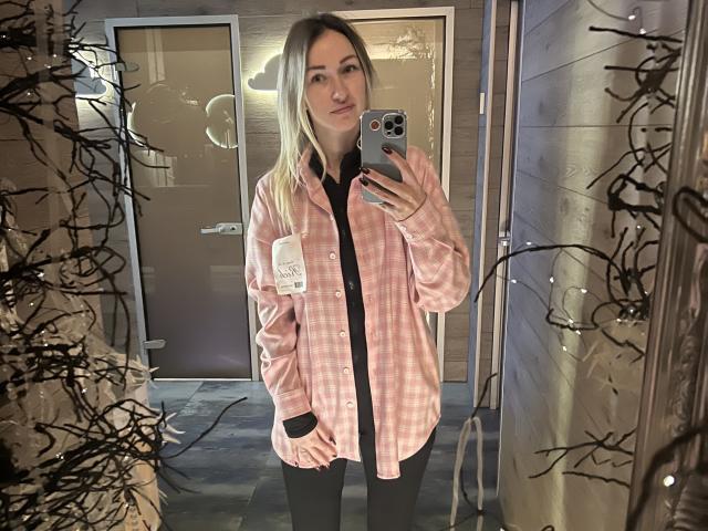 Lilitraya with blonde hair, pink plaid shirt, black shirt underneath, black pants, holding phone in mirror, wooden wall, black string lights, slight smile, medium-sized tits visible.