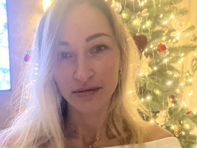 Lilitraya has light blonde hair, subtle makeup, and an off-shoulder white top, standing in front of a Christmas tree with warm lights and red ornaments.