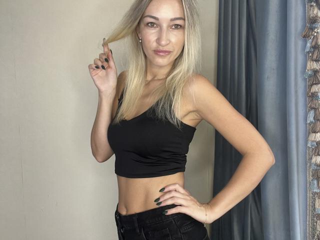 Lilitraya with blonde hair, black crop top, and black jeans, holding her hair, showing a small butt, and a confident smile, standing with one hand on her hip.