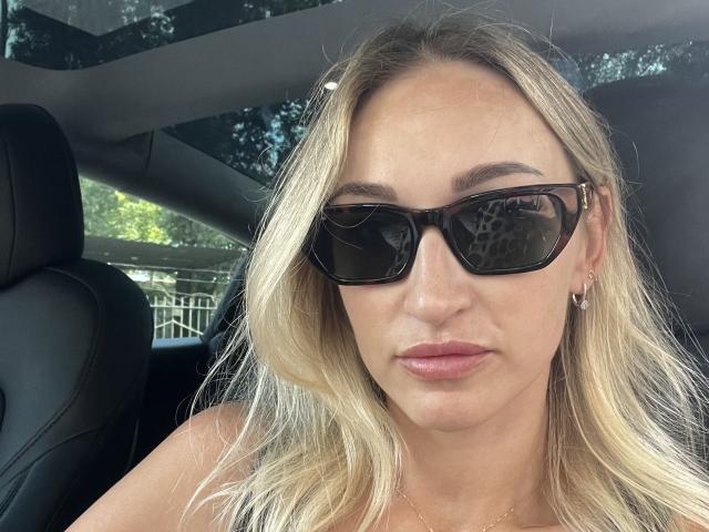 Lilitraya with blonde hair, wearing black sunglasses, subtle pink lipstick, and small earrings, looking straight at the camera, sitting in a car.