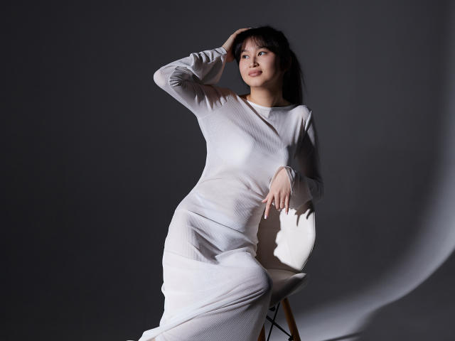 SheilaPass, mulher asiática, vestindo um vestido branco, sentada em uma cadeira branca, com a mão na cabeça, sombra na parede, luz suave, pose relaxada, sorriso discreto, cabelo preto longo, seios pequenos..