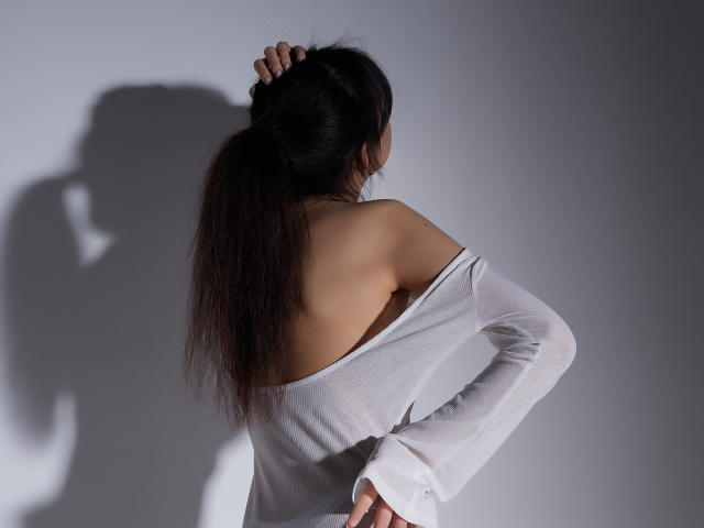 SheilaPass met kaal achterwerk, witte top met open schouder, donkere paardenstaart, hand in haar, schaduw op de muur, zacht licht, staand, rug naar de camera, slank, aantrekkelijk, subtiel sexy..