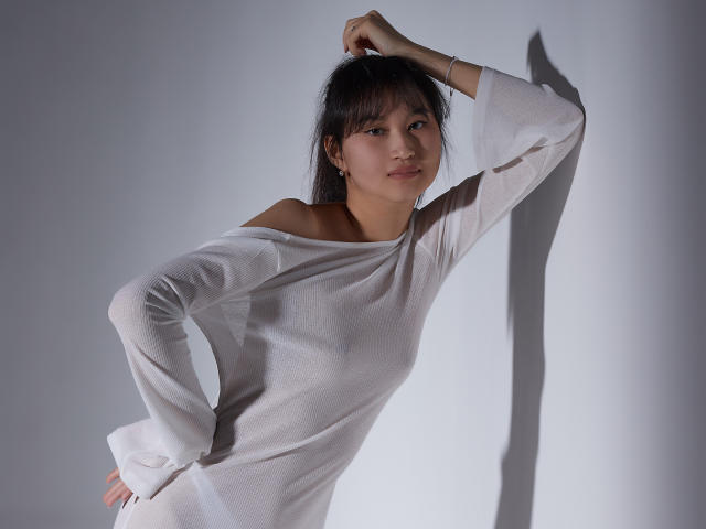 SheilaPass with dark hair, white off-shoulder shirt, hand in hair, subtle smile, standing, shadow on wall, soft light, relaxed pose, slightly bent, elegant, casual.