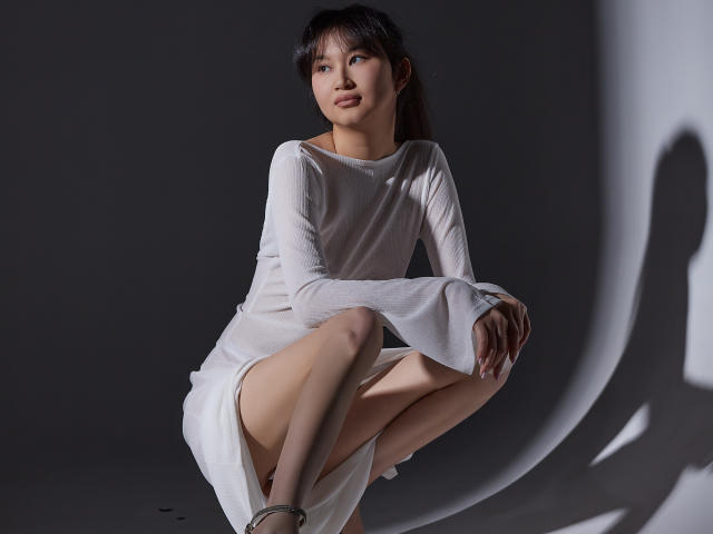 SheilaPass tiene la piel pálida, cabello negro y pechos pequeños. Está agachada con un vestido blanco, mostrando su trasero desnudo, con una sombra a la derecha. Lleva calcetines hasta los tobillos..