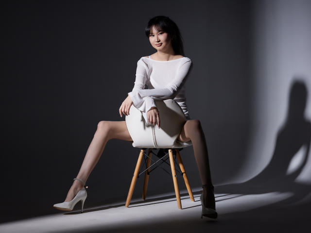 SheilaPass, assise sur une chaise, portant un haut blanc à manches longues, une mini-jupe blanche et des talons hauts blancs, avec les jambes écartées, montrant ses fesses nues..
