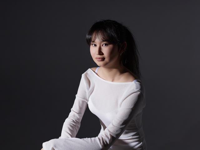 SheilaPass with small tits, wearing a white long-sleeve shirt, sitting against a dark gray background, looking to the side with a slight smile, black hair, and bangs.
