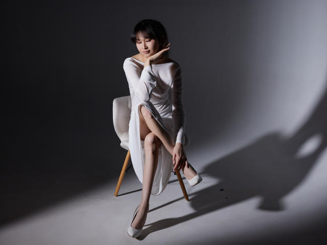 SheilaPass está sentada en una silla, vestida con un vestido blanco transparente, que deja ver su trasero desnudo a través de una abertura, con tacones blancos, y su sombra proyectada en el suelo..
