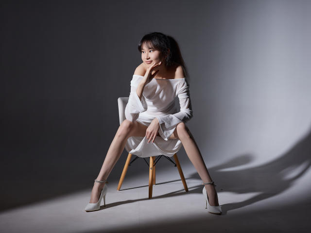 SheilaPass with tits slightly visible, sitting on a chair, legs spread, white off-shoulder dress, white high heels, shadow on gray wall, looking to the side, hand on chin.