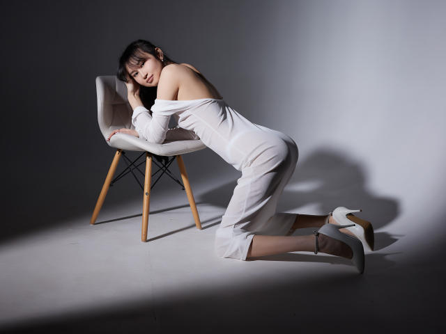SheilaPass está arrodillada con un vestido blanco, de corte asimétrico que deja ver sus pechos, frente a una silla, con tacones blancos, sombras en el suelo, mirando a la cámara, luz suave, pose sensual..