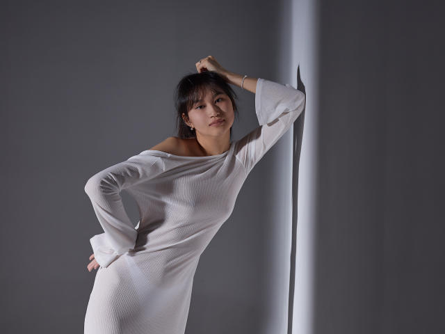 Sheila: Cabelo escuro, vestido branco com um ombro descoberto, mão na parede, outra mão no quadril, feixe de luz suave, olhando para a câmera, bumbum pequeno, corpo esguio, expressão calma..