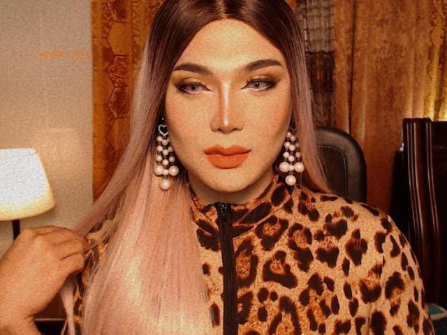 PattyHugeCock with long pinkish-brown hair, leopard print jacket, orange lipstick, and big pearl earrings, holding hair, looking at camera, sitting in a warm-toned room.