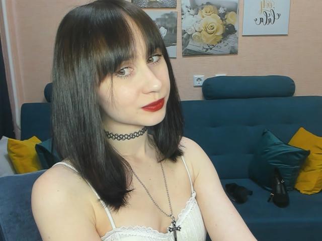 RebeccaRoger has black shoulder-length hair, red lipstick, and a black choker. She's wearing a white lace top, showing her small tits. She's sitting on a blue sofa.