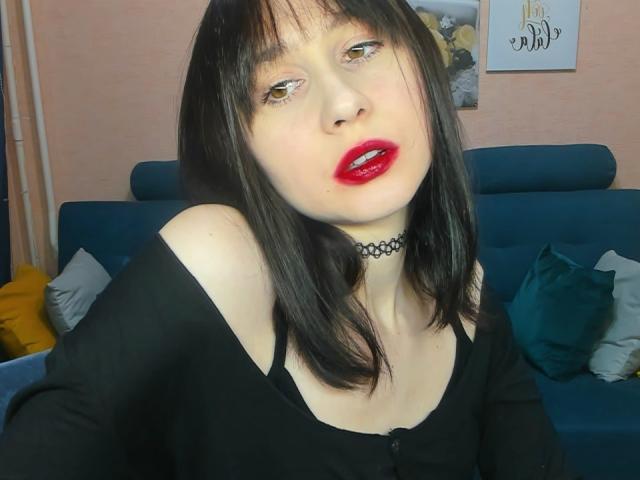 RebeccaRoger with pale skin, black hair, red lipstick, and a black choker, wearing a black off-shoulder top, showing her tits. She's on a blue sofa with pillows.