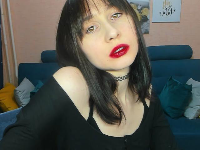 RebeccaRoger with pale skin, black hair, red lipstick, black choker, black top, and brown eyes, sitting on blue couch with yellow and green pillows, looking at camera.