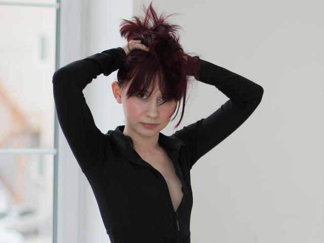 RebeccaRoger with hands in messy red hair, wearing a black zip-up, showing a bit of her tits, standing in a bright room, looking at the camera.