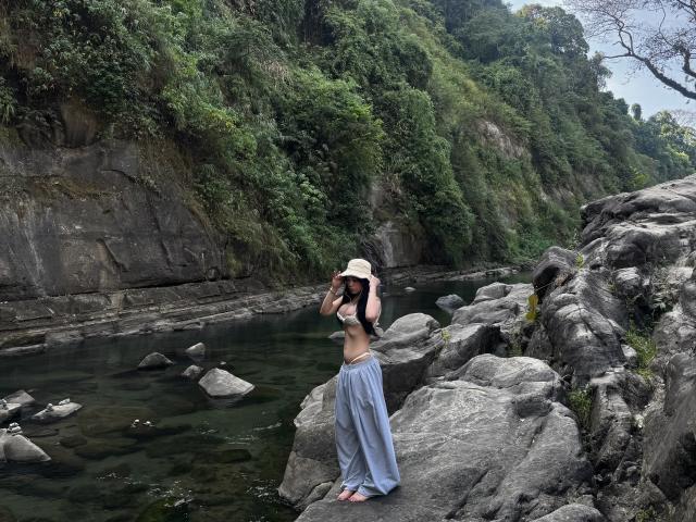 KassandraKate stands on rocky shore, wearing a beige hat, blue loose pants, and no top, revealing her small tits and slim waist. She's touching her hat, by a river.