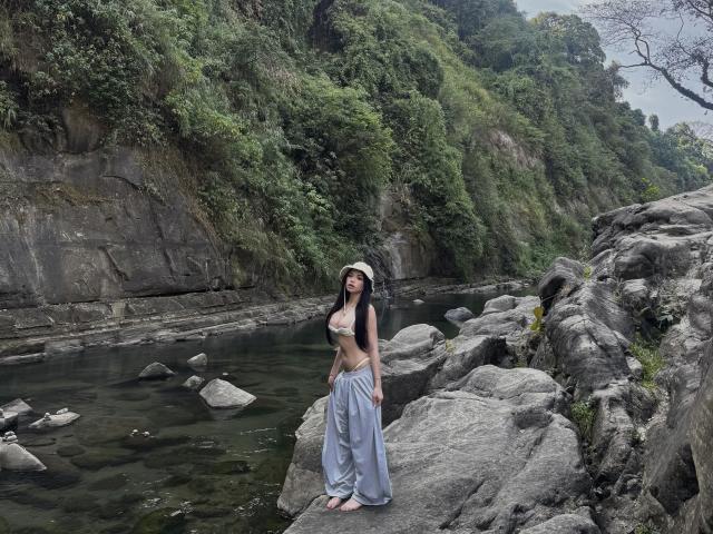 KassandraKate with small tits, wearing a white sunhat, beige bra, and loose gray pants, standing on rocks by a clear, rocky river with green forest in the background.
