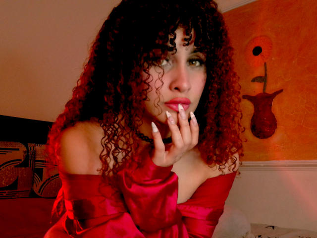 Amaia Escotet mit lockigem, rotem Haar, rotem Oberteil mit einem Schulter frei, schwarzem Choker, Hand in der Nähe des Mundes, Blick in die Kamera, rotes Licht, weiße Nägel, orangefarbene Wand mit abstrakter Kunst..