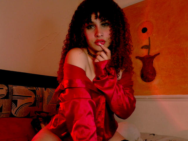 AmaiaEscotet with curly brown hair, red satin off-shoulder shirt, finger near red lips, sitting on bed, red lighting, wall with abstract horn-shaped decoration, seductive expression.
