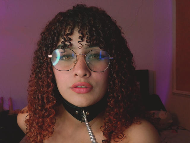 AmaiaEscotet with curly red-brown hair, wearing round glasses and a black collar with a silver chain, has pink lips and freckles, looking directly at the camera.