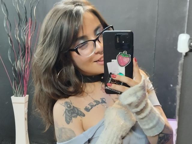 Amy, con gafas, cabello ondulado castaño, tatuajes en el pecho, top gris de corte asimétrico, mangas grises de felpa, tomando un selfie con una funda de teléfono con forma de fresa, collar negro, uñas rojas, sonriendo..