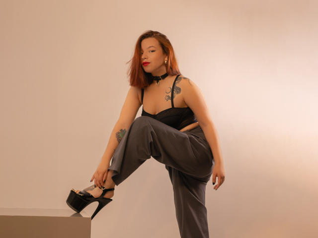 SabriinaMorgan, standing, one leg up on a box, wearing black strap top, gray pants, black high heels, red lipstick, tattoo on right shoulder, choker, looking at camera.